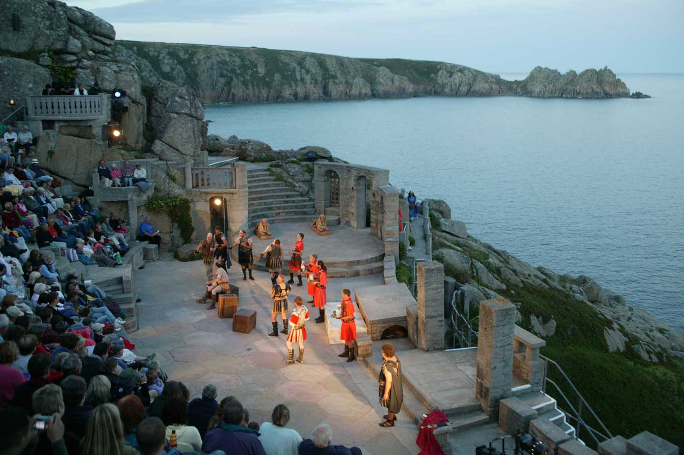 Open-air performance at the Minack Theatre with the audience watching against a backdrop of sea cliffs.