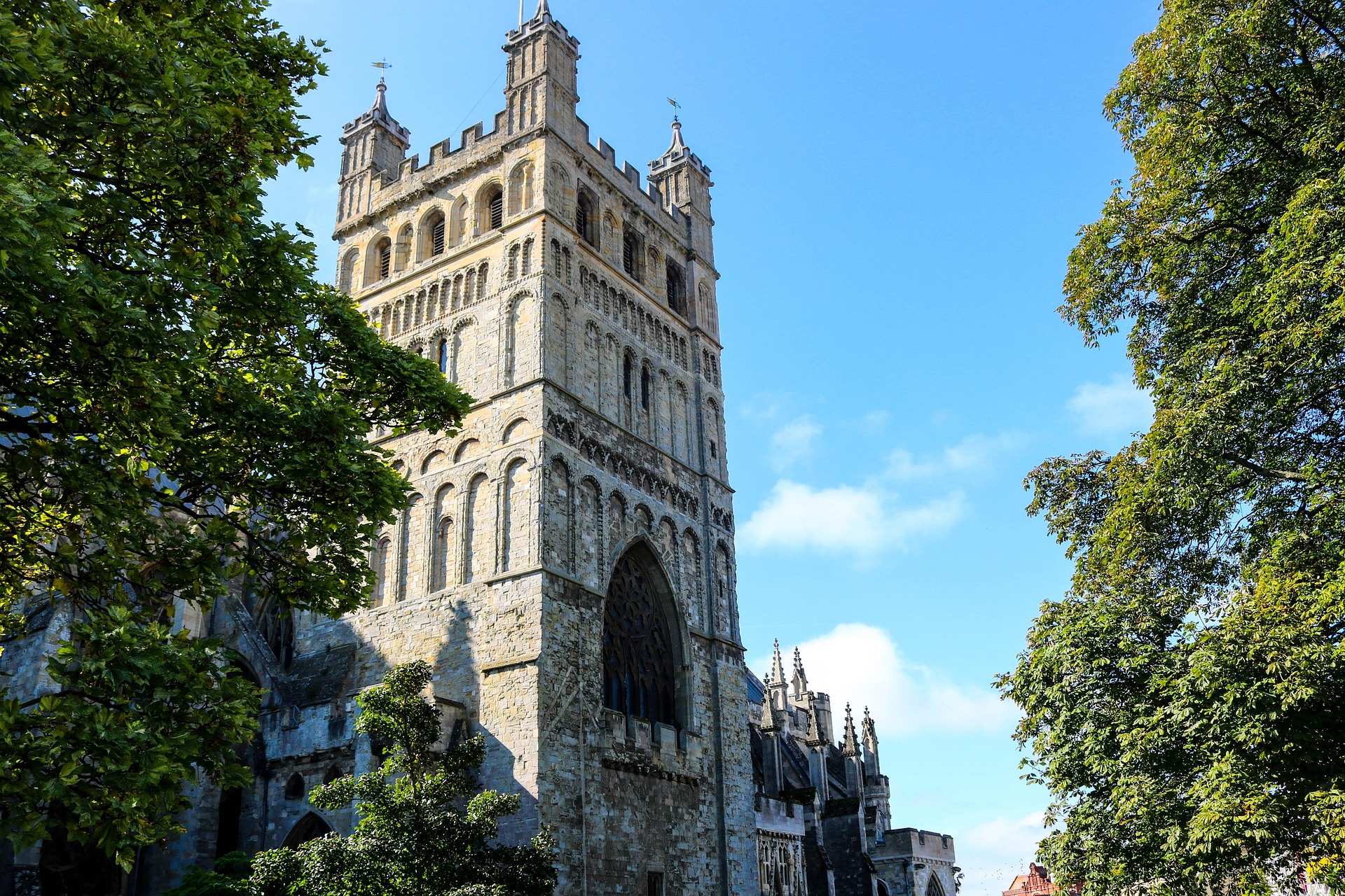 View of Exeter Cathedral’s tower rising above leafy trees against a bright blue sky.