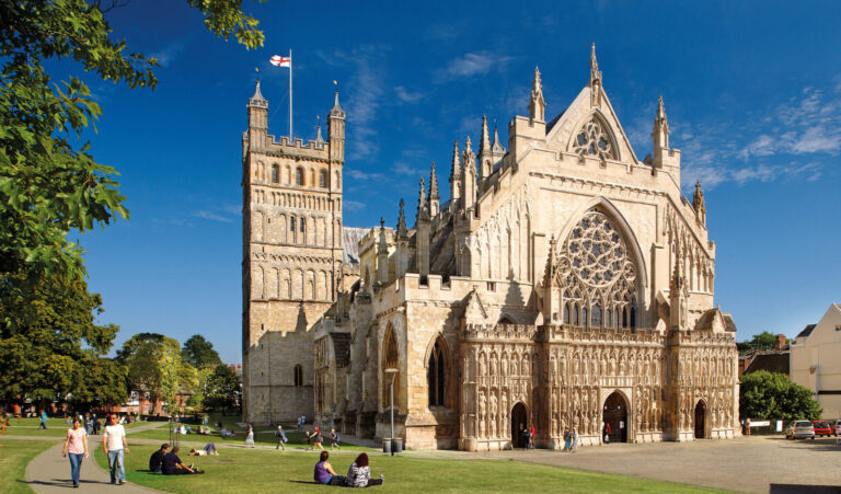 People relaxing and walking across the green in front of Exeter Cathedral under clear blue skies.