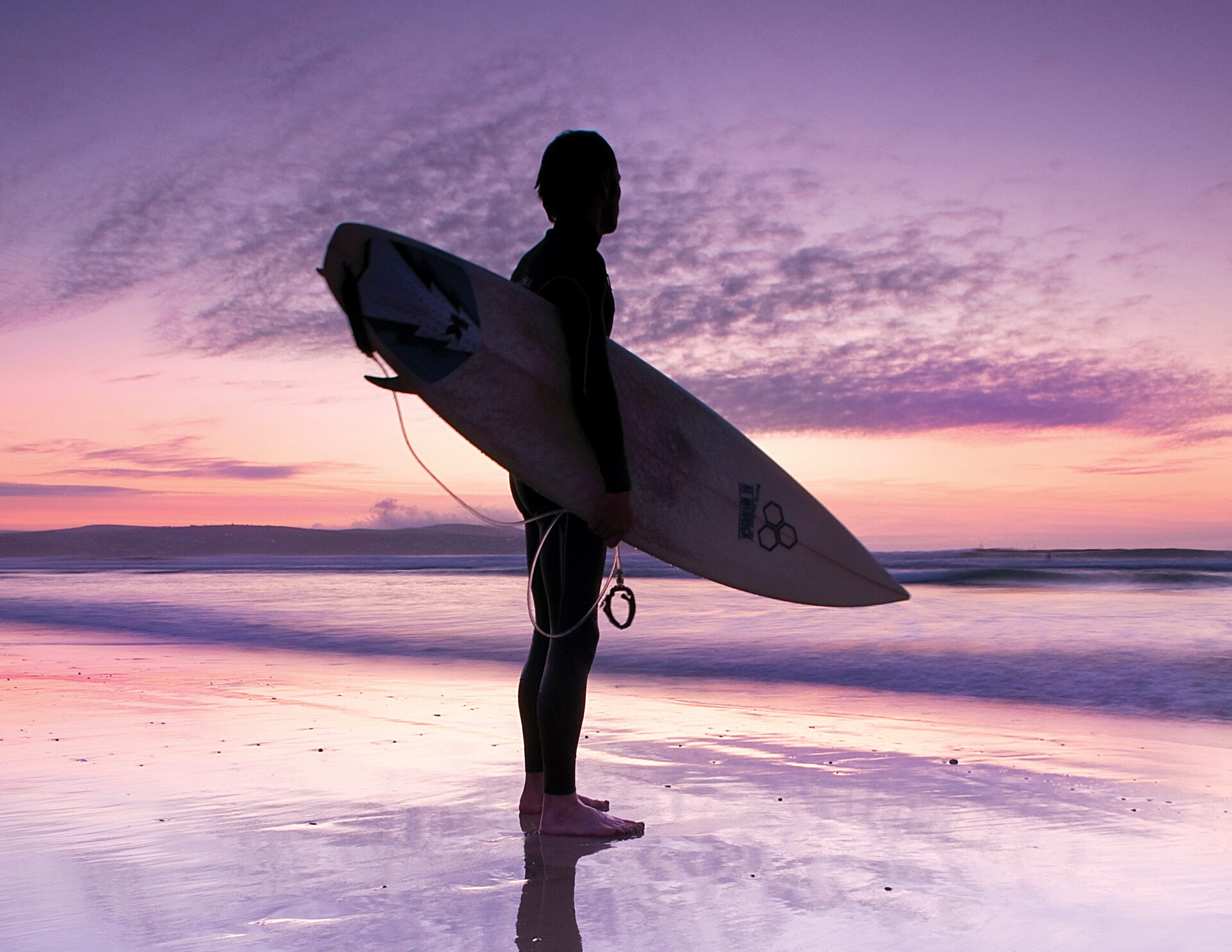 Silhouette of a surfer holding a surfboard at sunset on a Newquay beach.